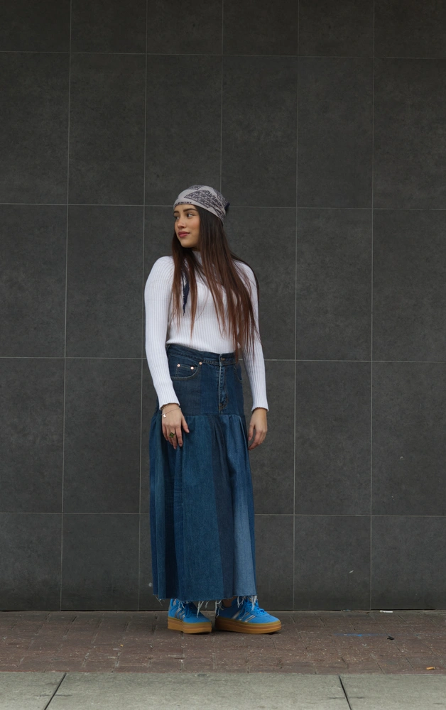 The 70s model looking with an averted gaze, standing in front of a grey tiled wall.
