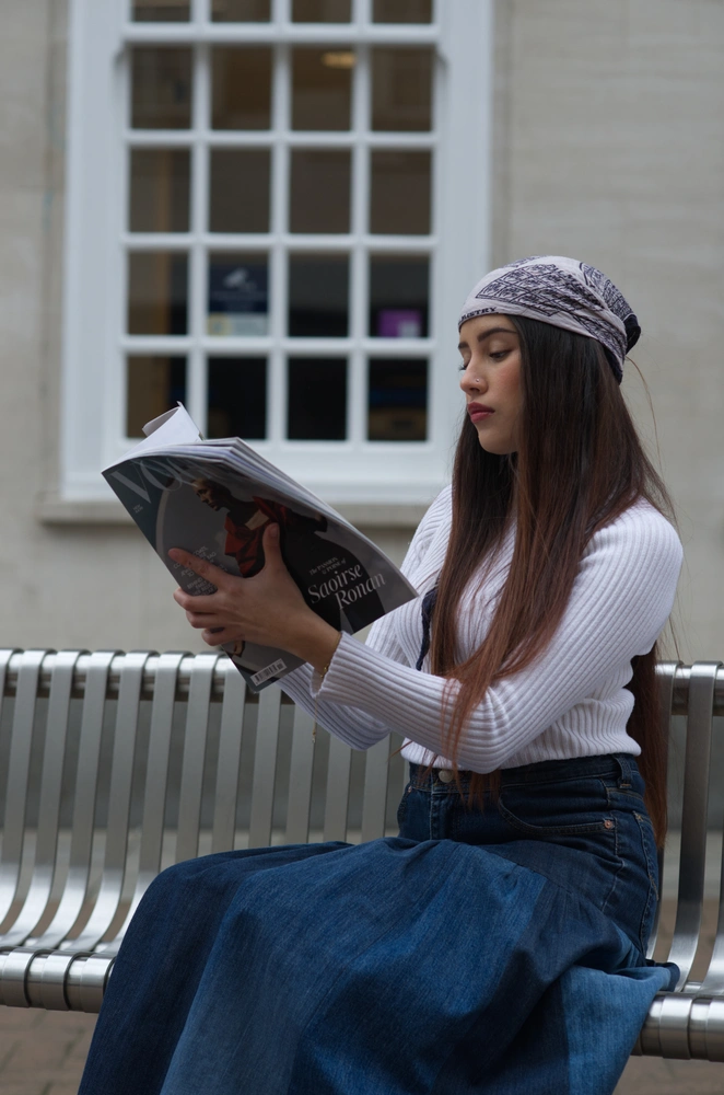 The 70s model sitting on a metal bench, posed reading a Vogue magazine.