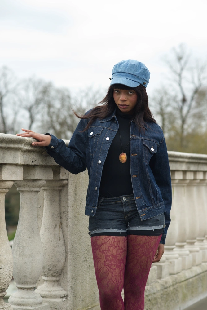 The 90s model standing, looking down towards the audience. The model is posed with her hand on the top of a bridge wall. Blured trees are in the distance, behind the model.